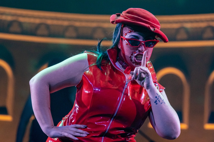A torso-up photo of Demon Derriere performing on stage wearing all red hat, mask, sunglasses, and pvc jacket. They are posing in a ‘shush’ position. The background has white frames on the wall with a shadowy orange and blue tinge.